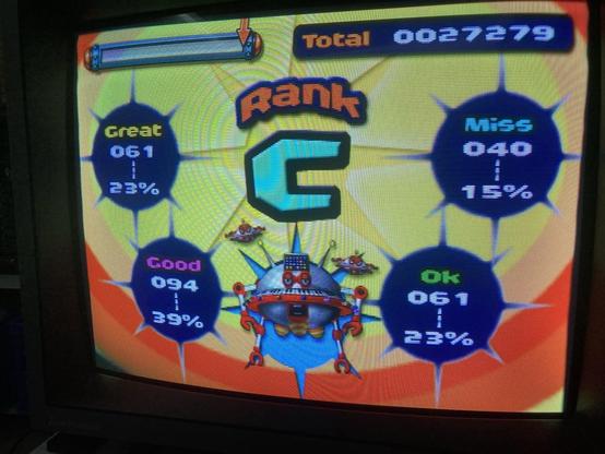 Results screen of Gitaroo Man after battle against the Flying-O boss fight.

On top edge of screen there a energy and points bars. Below them, center of screen sits a ranking letter for the battle. It is surrounded by an arc of four blue percentage score circles, and a picture of Flying-O character. The background picture consists of small yellow and green ring pattern, and wider red rings arcing towards bottom of screen.

Flying-O is sort of a sphere shaped flying robot with simple ball-jointed arms and legs. Two smaller battle robots hover to its left & right, both shaped more like flying saucers. 

Total game score is 27279 points, overall rank is C. The percentage scores in blue circles run between Great - Good - Ok - Miss and are 23% - 39% - 23% - 15% respectively.