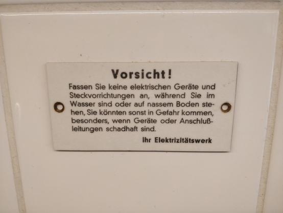 Schild auf einer weißen Kachel.
Text: Vorsicht! Fassen sie keine elektrischen Geräte und Steckvorrichtung an, während Sie im Wasser sind oder auf nassem Boden stehen. Sie können sonst in Gefahr kommen, besonders, wenn Geräte oder Anschlußleitungen schadhaft sind. Ihr Elektrizitätswerk