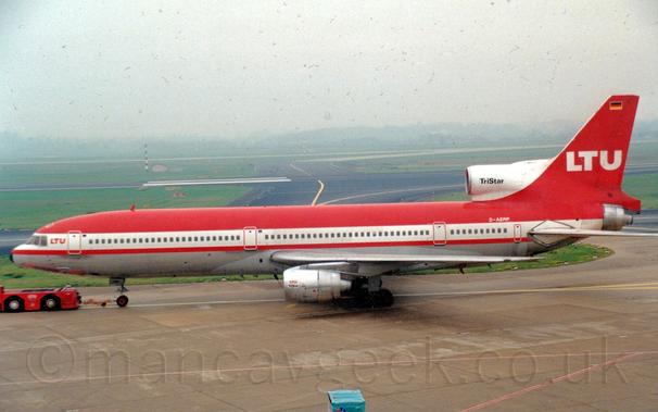 Side view of a red and white, 3 engined jet airliner being towed from right to left by a low-slung, red tug attached to the nose-wheel by a along yellow pole.
The plane has red upper surfaces with a metallic grey belly, with a thick white stripe running along the body covering the cockpit and passenger cabin windows.
There are small, red, "LTU" titles on the upper forward fuselage, between the rear of the cockpit and the forward door.
The red tail has large white "LTU" titles in the middle, with a small German flag at the top.
The under-wing engine pods are a bare metal, with small, red "LTU" titles and black "TriStar" text next to the intake.
The centre engine intake in front of the tail is white, with black "TriStar" text,
Grey concrete apron fills the foreground, with green grass and black taxiways and a runway vanishing into mist in the background.