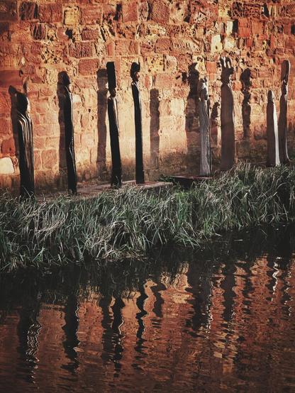 The image shows a row of 8 figures standing before a brick wall. In the foreground is a body of water reflecting the figures and the wall. Green grass grows between the figures and the water. The figures themselves vary in appearance; some are dark and columnar, while others are lighter in color and have more distinct, carved shapes. Shadows are cast to the left of the figures, suggesting a light source from the right. The wall behind the figures is constructed of reddish-brown bricks.