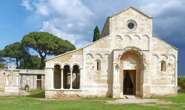 The image features a decorative stone building, likely a church or a historical structure, characterized by its Romanesque architectural style. Key elements include arched windows, a round window at the top, and a portico with arches. The surrounding area has grassy land and trees, suggesting a peaceful outdoor setting. The sky appears cloudy, enhancing the historical ambiance of the structure.

Image Credits: Wikimedia / Holger Uwe Schmitt / CC BY-SA 4.0
