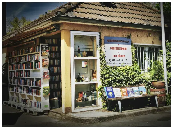 Ehemaliges Pforten- und Empfangshäuschen einer Brauerei, umfunktioniert zu einem Showroom für Artikel aus Entrümpelungen und Haushaltsauflösungen, vorwiegend Bücher. Das Haus wurde 'über Eck' aufgenommen, auf der linken Seite ein großes weißes Bücherregal, daneben der Eingang mit einem weiteren Regal. Auf der rechten HauswandEine in dei Wand eingelassene wandhohe Vitrine, daneben eine großes Reklameschild, rechts daneben ein großes vergittertes Fenster. Davor eine Bank, an der Rückenlehne sind weitere Bücher aufgestellt, auf der Sitzfläche werden ebenfalls Bücher ausgestellt und zum Verkauf angeboten. Farbgebung, Bildaufbau und Inhalt erinnern an Fotos der 60er/70er Jahre.