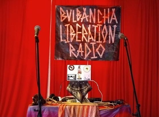 A Bvlbancha Liberation Radio handmade sign on stage in front of a red curtain, with a DJ setup on a laptop, and microphones on a table in the center.