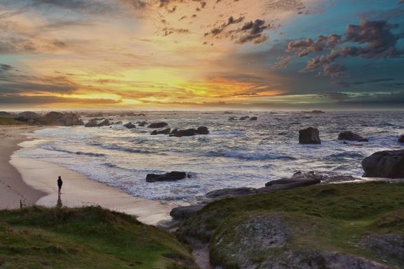 A serene beach scene at sunset, featuring gentle waves lapping against the shore, rocky formations, and a silhouette of a person standing near the water's edge. The sky is filled with vibrant colors, ranging from orange to blue, and scattered clouds.