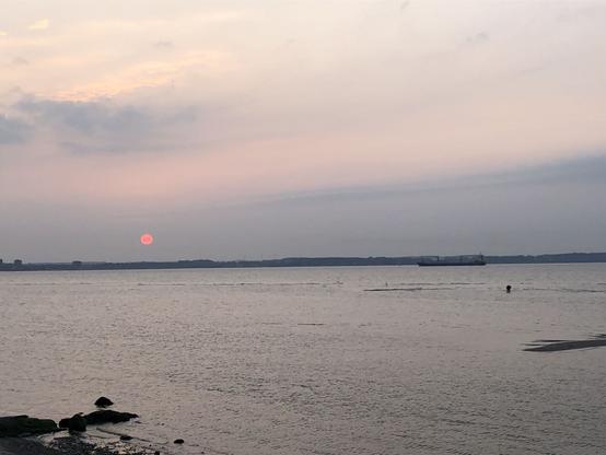 Eine kleine rosarote Sonne im hellblau des Himmels über dem Uferstreifen gegenüber. Rosa Streifen am Himmel und im Wasser gespiegelt. Ein Frachtschiff in Richtung Förde.