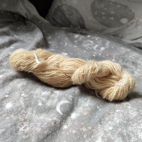 A photo of a skein of handspun 2-ply yarn. The yarn is a pale peachy colour and is twisted around itself, tied with white thread. The yarn is sat on top of a pale grey-blue duvet cover with a moon and stars pattern.