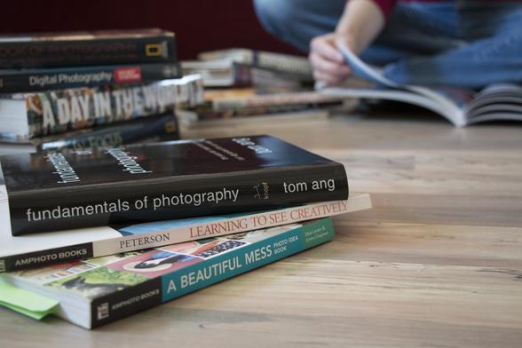 A stack of books about photography sits in the foreground, with another stack behind. Slightly out-of focus in the background, we see a portion of a woman sitting cross-legged on the floor, surrounded by the books, turning the page of the book she is reading.
