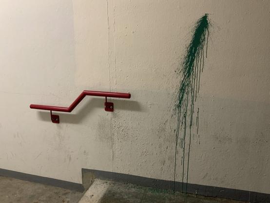 Ein Innenraumfoto zeigt eine kahle, beigenfarbene Wand mit einem roten, eckig geformten Handlauf, der an der linken Wand befestigt ist. Rechts daneben ist ein auffälliger grüner Farbfleck, der aussieht, als sei flüssige Farbe von einem Punkt oben an der Wand herabgelaufen und getropft. Der Boden ist aus rohem Beton und an der Wand sind Schmutz- und Wasserflecken zu sehen.
A photo of an indoor stairwell corner shows a beige wall with a red, angular handrail mounted to the left. To the right of the handrail, a streak of dark green paint appears to have been splashed or spilled from a high point, running down the wall in long, uneven drips. The concrete floor is rough and stained, and a dark zig-zag line follows the base of the wall, adding to the scene’s gritty, unpolished character.