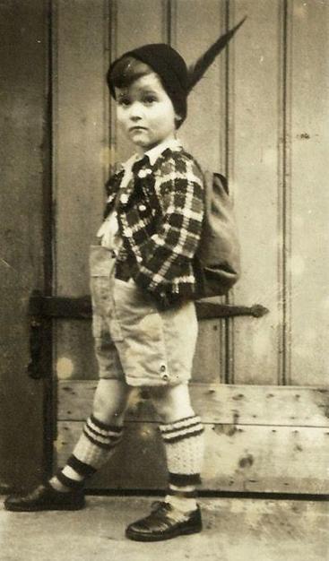 Photo of a young boy - the whole silhouette. He is standing against a wall of boards. He is wearing a plaid shirt, short shorts, thick knee socks and a hat with a feather.