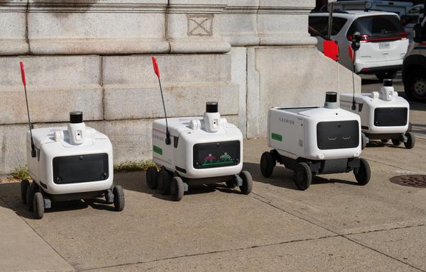 Four delivery robots (looking like picnic coolers with wheels below, displays for fake eyes in front, and sensors and flag masts atop) are parked in a row.
