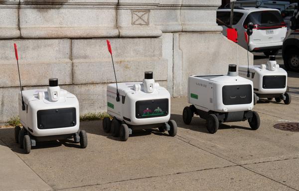 Four delivery robots (looking like picnic coolers with wheels below, displays for fake eyes in front, and sensors and flag masts atop) are parked in a row.