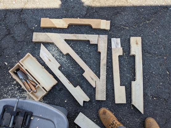 An arrangement of pieces of wood from old industrial pallets on a tarmac surface with scattered wood shavings and a wooden box of wood chisels with wooden handles are arrayed in feont of a grey plastic box with a 4 inch drill vise on top of it and my boots in the shade of the Vulpibus. The pieces of wood are in various stages of being chiselled and cut to shape, using the forklift gaps as arches to spread weight and as notches to rest the diagonals of the triangular bracket shapes on.