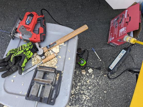 An arrangement of tools on and next to a grey plastic box, with a pair of work gloves at left in front of a jigsae, a drill vise in front of a hammer and chisel, and on the ground a tape measure (retracted), cables including an EV charger, pens amd the box for the jigsaw. Sawdust and shavings are scattered around.