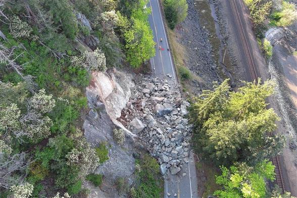 Aerial view looking down on a rockslide. Rocks slide of the hill on the the left side of the road and are entirely blocking both lanes. To the right of the road, further down the steep incline is a railroad track running parallel to the road.
