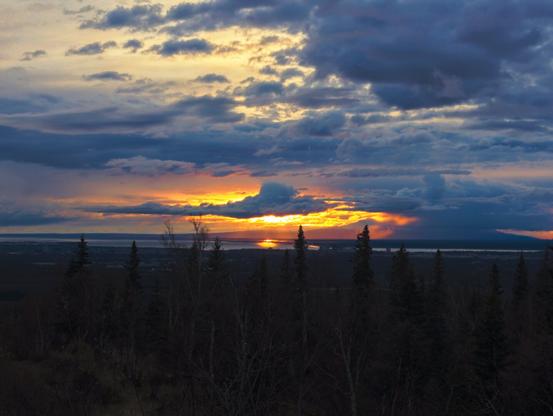 Sunset from the hillside above Anchorage, Alaska, looking towards Mount Susitna.  Some clouds, a bit puffy are in the sky.  One, rimmed with yellow and orange light, blocks the sun which is just above the horizon.  The sum glints off the Kinik arm in the distance.  The shadowy shapes of spruce trees are in the forground.