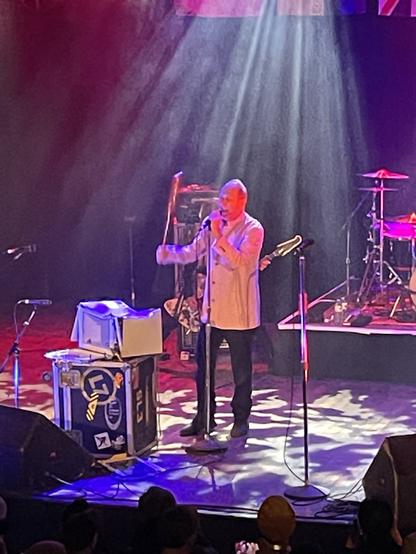 A photo shows an older-looking man in dark trousers and a white jacket. He is Jon King, the lead singer of the Gang of Four, and he’s raising a baseball bat over a badly damaged microwave as well as singing into a mike on a stand.