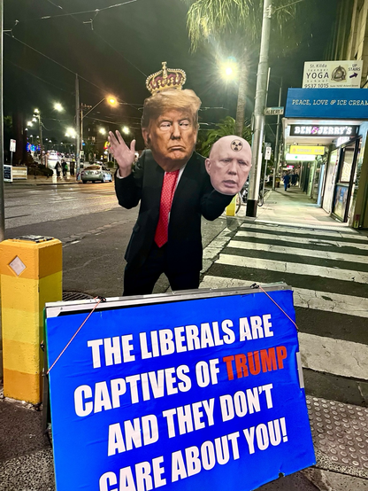 A man with two heads, one is Trump the other is Dutton, standing in a street holding a sign that says “The liberals are captives of Trump and they don’t care about you.”