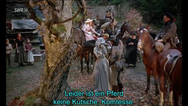 Description: The image shows a group of people riding horses down a dirt road, with a tree on the left side and a few people standing on the ground. In the background, there are many trees, and at the bottom of the image there is some text which reads "Leider ist ein Pferd keine Kutsche, komtesse".

Disclaimer: This image description was automatically generated and may not be entirely accurate.