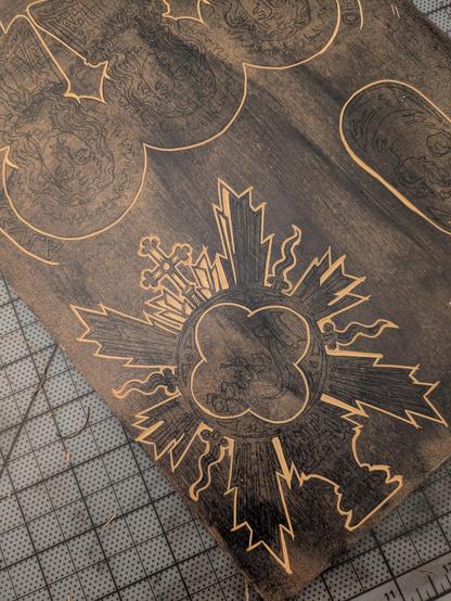 A linoleum block laying on a cutting mat. Designs are partially carved into it. The carving furthest along represents a "sunburst"-shaped monstrance with the word "Relics" (backwards for printing) in the center.