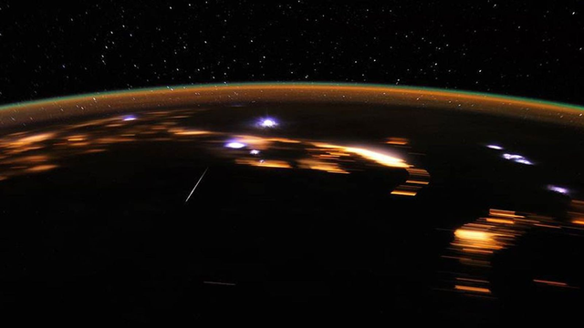 Image taken from the International Space Station showing the Lyrid meteor shower from above. The Earth's atmosphere is a thin line across the image. Above are stars on black sky. Below several bright, burning meteors are visible.