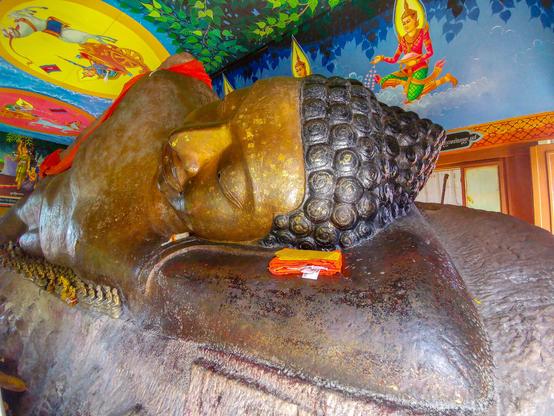 Photo shows the top part and head of a reclining Buddha-statue. The figure is resting on its left arm with eyes closed. The stone crop it is a part of and carved out of is clearly visible. A small offer of rolled together clothing with some money notes is placed as a headrest. The photo shows the whole figure ten meter down to its feet in perspective from the top head. From the statues armpit to its leg there is a narrow shelf between statue and stone, where a range of smaller Buddha statues in sitting pose and different colours are placed. The roof and walls of the building built over the statue is richly and colourful decorated with scenes from the first Buddhas life.