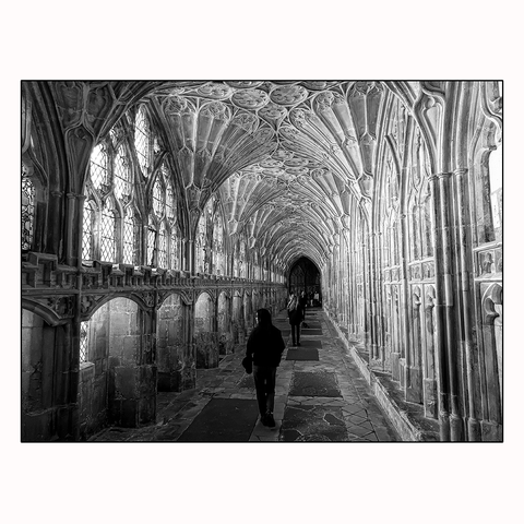 Several people are walking down a cloister corridor with 14th-century medieval fan vaulted ceilings and stained glass windows. The floor is made up of polished slabs of stone, some of which are horizontal tombstones.