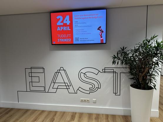Image of a net presenter screen in an office hallway, next to a potted plant. The net presenter shows a slide to announce today's strike, with the program.
