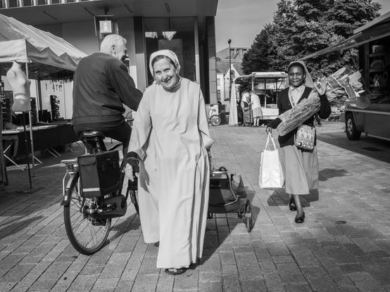 The image depicts a lively outdoor scene in Bruges, Belgium. In the foreground, three individuals are visible. On the left, an elderly man is seated on a bicycle with a cargo box attached, engaging in conversation with a woman in the center. She is dressed in a long, light-colored robe and headscarf, smiling as she walks. To the right, another woman, also wearing a headscarf, carries a bouquet of flowers and a white shopping bag, walking away from the camera. The background features market stalls, a food truck, and a building with a canopy, suggesting a bustling environment. The scene is captured in black and white, emphasizing the textures and contrasts.