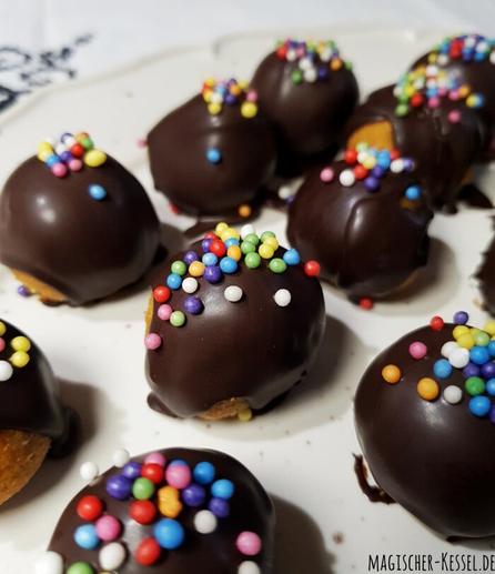 Weiße Tischdecke mit schwarzer, geometrischer Stickerei, darauf eine ovale Servierplatte mit geschwungenem Rand aus hell glasiertem Steingut. Auf der Platte sind kleine Kuchenpralinen zu sehen. Stellenweise erkennt eins die möhrenfarbene Füllung. Sie sind mit dunkler Schokolade glasiert und mit dicken, bunten Zuckerperlen verziert.