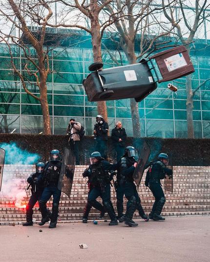 Small group of riot cops bunched up at the bottom of some concrete steps in a public space with a flare burning behind them. They are beginning to scurry as a large plastic residential trash can is flying through the air, about to hit them.