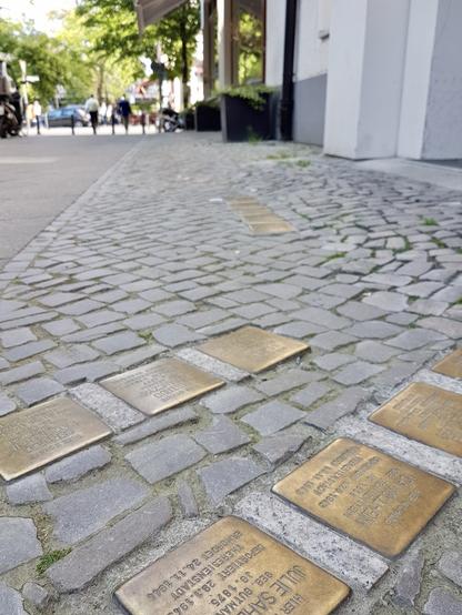 several brass memorial plaques, known as "Stolpersteine" or "stumbling stones," embedded in a cobblestone sidewalk in Berlin. These small brass plaques are flush with the cobblestones and contain engraved text.

Stolpersteine are Holocaust memorials created by German artist Gunter Demnig to commemorate victims of Nazi persecution. Each stone is typically placed in front of the last known residence or workplace of the victim before their deportation or murder. The brass plates usually contain the person's name, year of birth, date of arrest, fate (such as deportation or murder), and date and place of death if known.

In the background of the image, there's a street with some pedestrians walking, trees providing shade, and storefronts. The cobblestone pavement has some grass growing between the stones, and there appear to be at least 6-7 Stolpersteine visible in this particular location, suggesting that multiple victims may have lived or worked at this address.

These memorial stones serve as a decentralised monument throughout Europe, creating a tangible reminder of the Holocaust integrated into the everyday urban landscape.