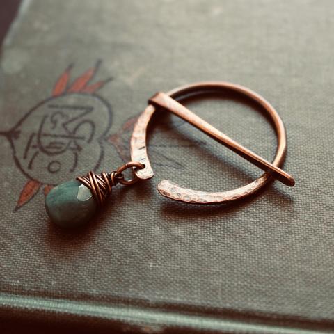 A photo of a circle-shaped, hand-forged copper brooch/pin resting on a vintage green hardcover book. The brooch has a green moss agate stone attached with wire wrapping, dangling near the bottom opening of the circle.
