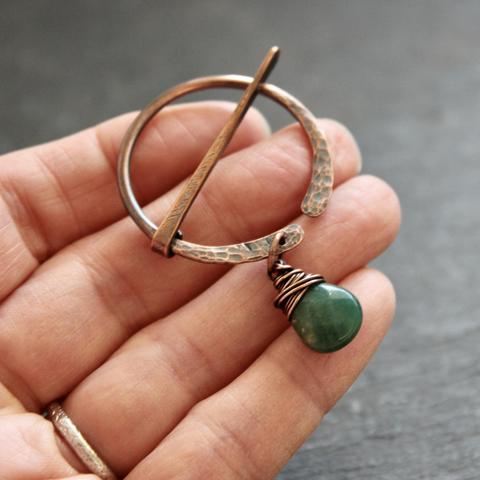 A photo of a circle-shaped, hand-forged copper brooch/pin in the palm of a hand. The surface of the brooch in hammered and patina'ed; the circle portion of the brooch has a green moss agate stone dangling from it with wire wrapping.