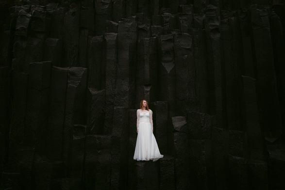 The image shows a bride at her elopement in Iceland standing gracefully on a formation of tall, dark basalt columns in Iceland. She wears a flowing white wedding gown with long lace sleeves, which creates a striking contrast against the rough, geometric black rock behind her. The basalt columns rise vertically, creating a dramatic and textured natural backdrop. The bride’s calm and poised presence adds a soft, elegant touch to the rugged landscape, capturing a moment of serene beauty and natural grandeur. An image taken by Hamburg based European wedding and elopement photographer Sturmsucht.