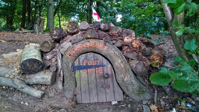 The image shows a whimsical scene of 'Peter Rabbit's Post Office'. The post office is built into a small mound, appearing like a hobbit-style dwelling. The entrance is an arched wooden door with the words 'PETER RABBIT'S' in orange lettering above the door and 'POST OFFICE' written below. A black hinge and a round doorknob are visible on the door. The structure is constructed from logs and branches, giving it a rustic, woodland appearance. A small English flag is seen in the background. The surroundings are lush with greenery and trees, suggesting a forest setting.