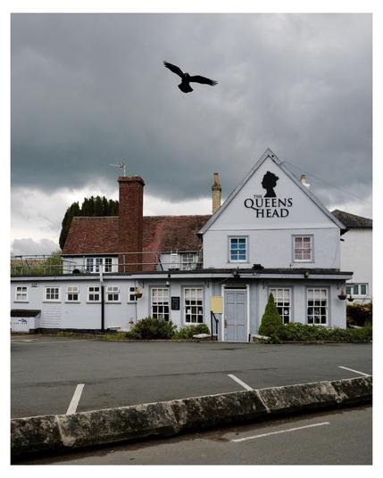 The image shows a two-story building called "The Queens Head," which appears to be a pub, given the traditional architectural style and signage. It has a clean white facade with blue trims around the windows and doors. The building is contrasted by a gloomy, cloudy sky, which adds a dramatic atmosphere. A bird, possibly a crow or raven, is flying in the sky above the building, enhancing the moody feel of the scene.