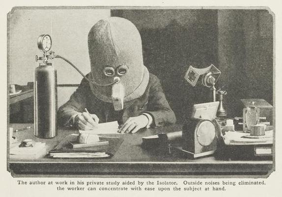 A black-and-white photograph showing a man seated at a desk, wearing a large, cylindrical helmet-like device called "The Isolator", designed to eliminate outside noise. The device covers his entire head and includes glass eye pieces and a breathing tube connected to an oxygen tank. He is intently writing with pen and paper, surrounded by early 20th-century office equipment. The scene is described in the caption below the image: "The author at work in his private study aided by the Isolator. Outside noises being eliminated, the worker can concentrate with ease upon the subject at hand."