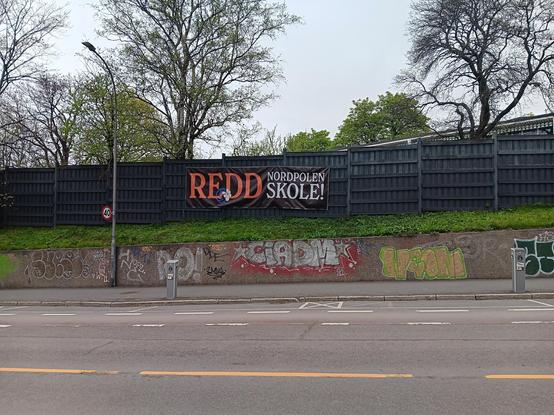 A sign displaying, "REDD NORDPOLEN SKOLE" [Save Nordpolen school] at the side of the road, attached to a dark wooden fence.