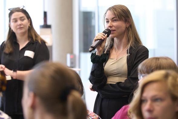 Eine junge Frau mit Mikrofon spricht, sie steht im Publikum einer Veranstaltung. Mehrere andere Frauen sind zu sehen.