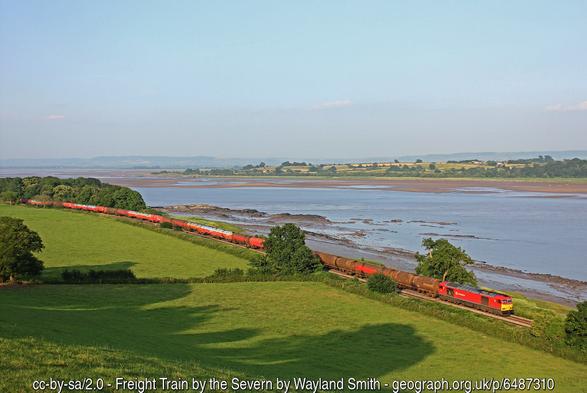 From high ground a view of a green plain bordering a wide fiver.  A long freight train is passing along the riverbank