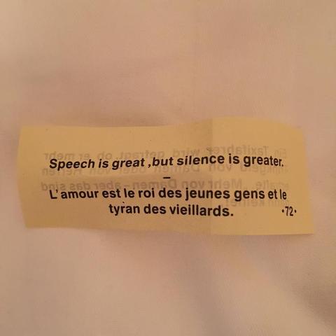 "A yellow fortune cookie with French text on it, placed on a white surface, quoting 'Speech is great, but silence is greater.' The mood appears to be serious or contemplative."