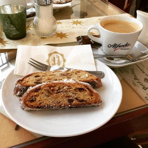 "This picture shows a plate of bread with nuts on top, placed next to a cup of coffee on a dining table covered with a tablecloth."