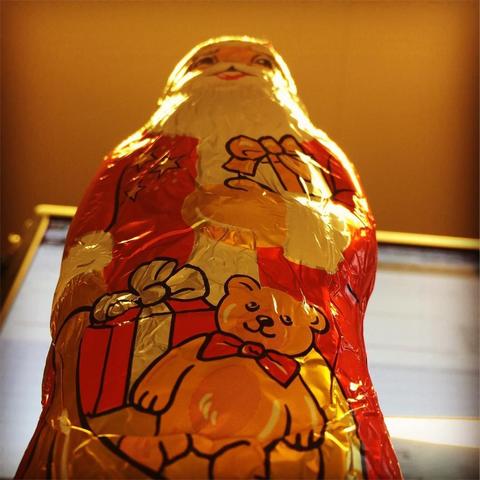"This picture shows a chocolate candy bar wrapped as Santa Claus, complete with reindeer antlers, a red suit, and white trimmed shoes, positioned in front of a Christmas tree."