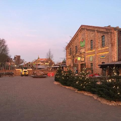 "This picture shows an outdoor holiday market with festive decorations and a building with signs, set against a dusky sky during what appears to be winter."