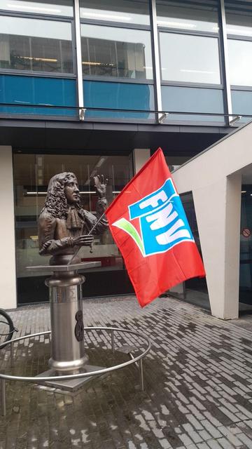 A sculpture of Christiaan Huygens in front of a building. He is holding a lens up in his left hand, looking through it. Somebody put a red-and-blue FNV flag in his right hand.