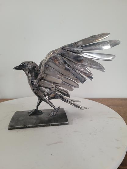 Sur la photo une toute petite corneille en acier est en train de marcher. Elle a les ailes déployées. Les ailes sont composées de manches d'ustensiles. C'est tout ce qu'on peut encore identifier.