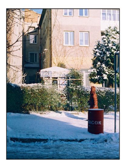 The image depicts a snowy urban scene. In the foreground, there is a red barrel with the word "NICOK" written on it, standing on a snow-covered ground. A person in a brown coat is walking past the barrel. Behind the barrel, a hedge covered in snow is visible, with a small white structure, possibly a kiosk, partially obscured by the hedge. The background features a multi-story building with beige walls and several windows, some of which have white frames. A tree with snow on its branches is also present, adding to the wintry atmosphere. The sky is clear and blue, indicating a sunny day. The scene is framed by bare tree branches on the left, contributing to the overall winter setting.