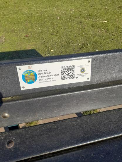 Close up of the back of a bench with a plaque on it which reads, “This is a Hello Bench. A place to sit, chat and connect.” Beside the text is a QR code.