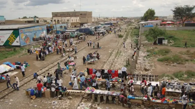 A bustling African outdoor market scene set near railway tracks, featuring numerous vendors selling various goods. The area is populated with people browsing and purchasing items, while others transport goods. Colourful clothing and merchandise are displayed on makeshift tables alongside the tracks, with buildings and buses visible in the background. The sky is overcast.