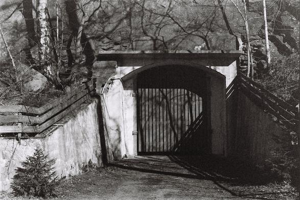 A grainy and slightly faded black-and-white photograph of a large door or gate going straight into a mountainside. The door is surrounded by a simple concrete structure big enough only to just fit the door and prevent dirt from falling onto the driveway in front. The structure appears to give no indication of what lies inside.